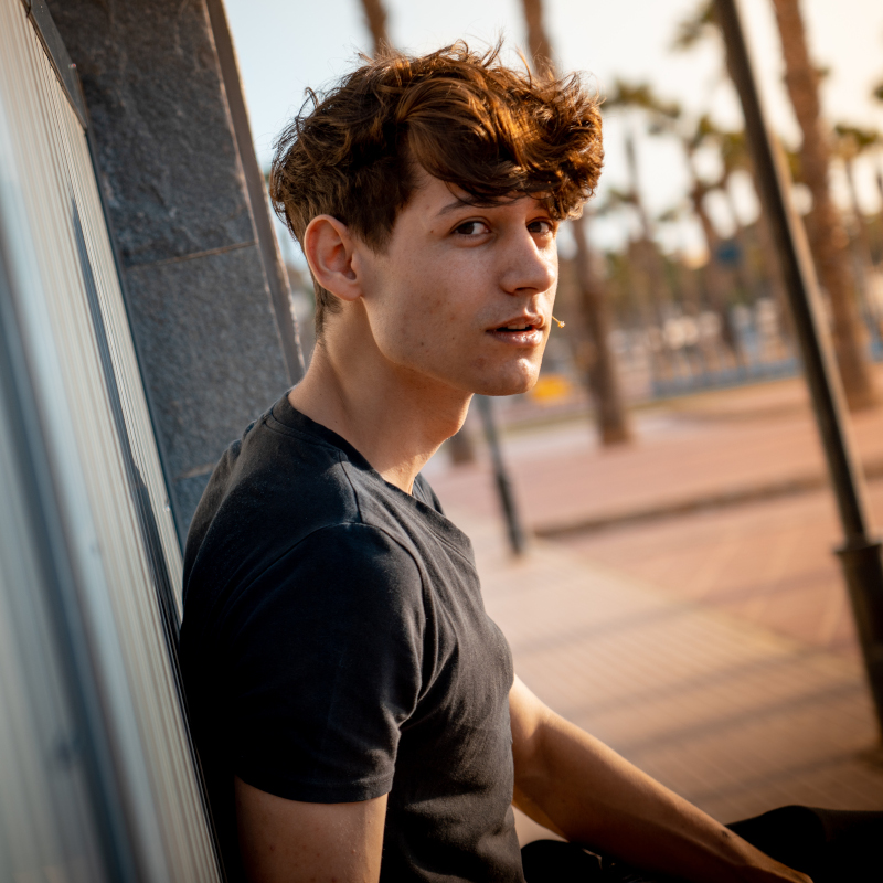Foto de perfil de un hombre con pelo corto y una camiseta negra ajustada donde aparece una playa con palmeras de fondo.